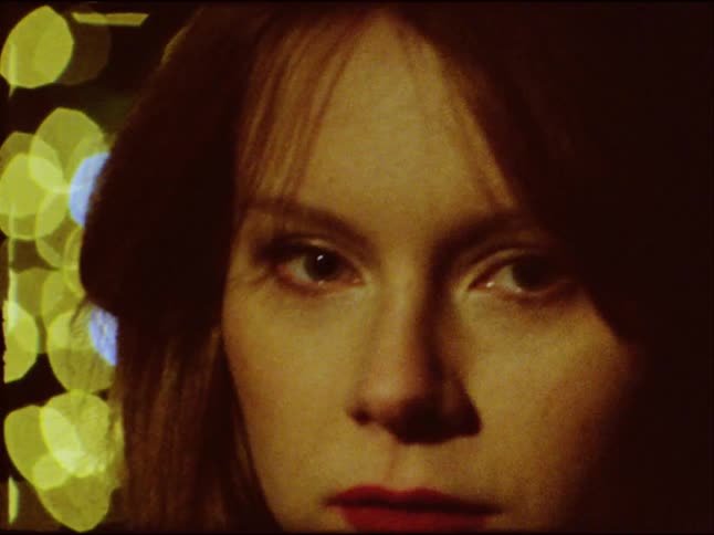 A vintage Super 8mm film shows a close-up of a mysterious woman's face as she stares intensely against a background of sparkling lights