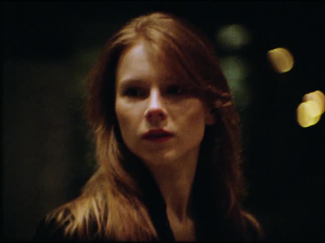 Vintage Super 8 film captures a mysterious woman with long hair looking over her shoulder on a dark city street at night