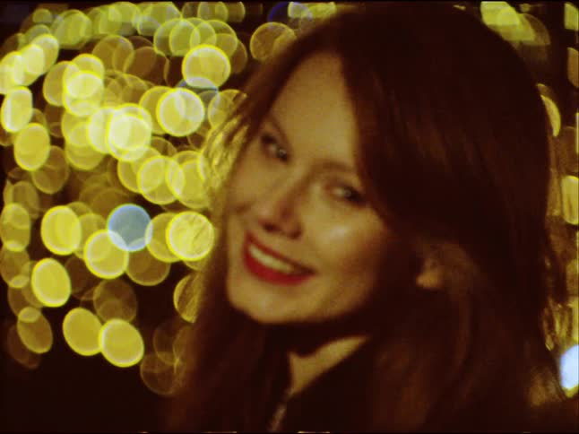 A vintage film style clip of a happy young woman smiling and laughing in front of a warm, golden bokeh light background at night