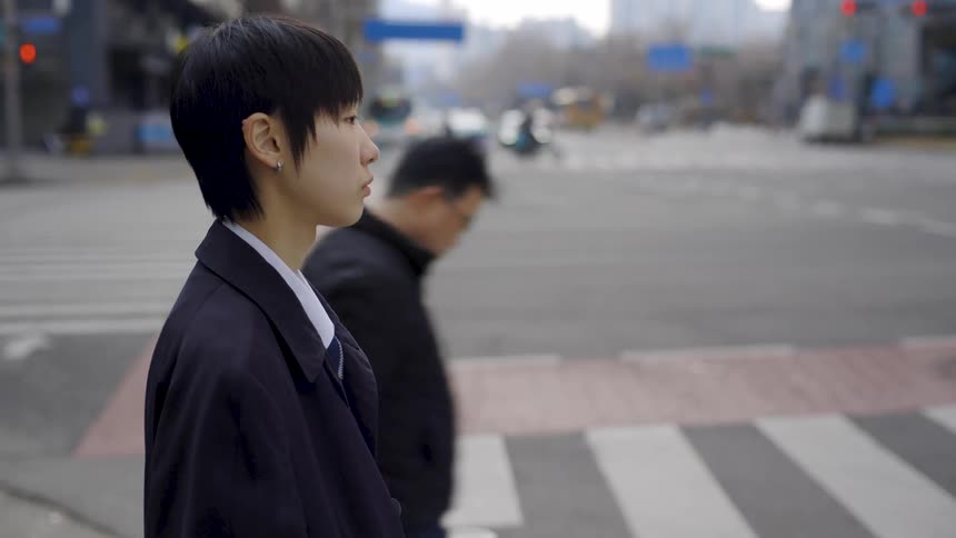 A pensive young person with short hair waits patiently at a busy city crosswalk in slow motion