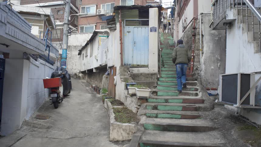 A man wearing a green jacket walks up a steep, weathered staircase in a narrow alley of an old residential neighborhood