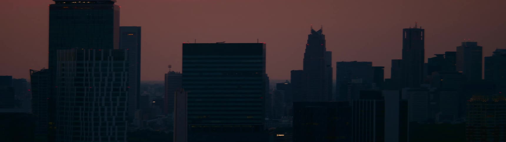 A hazy red sky glows over a city skyline at dusk as building lights begin to turn on