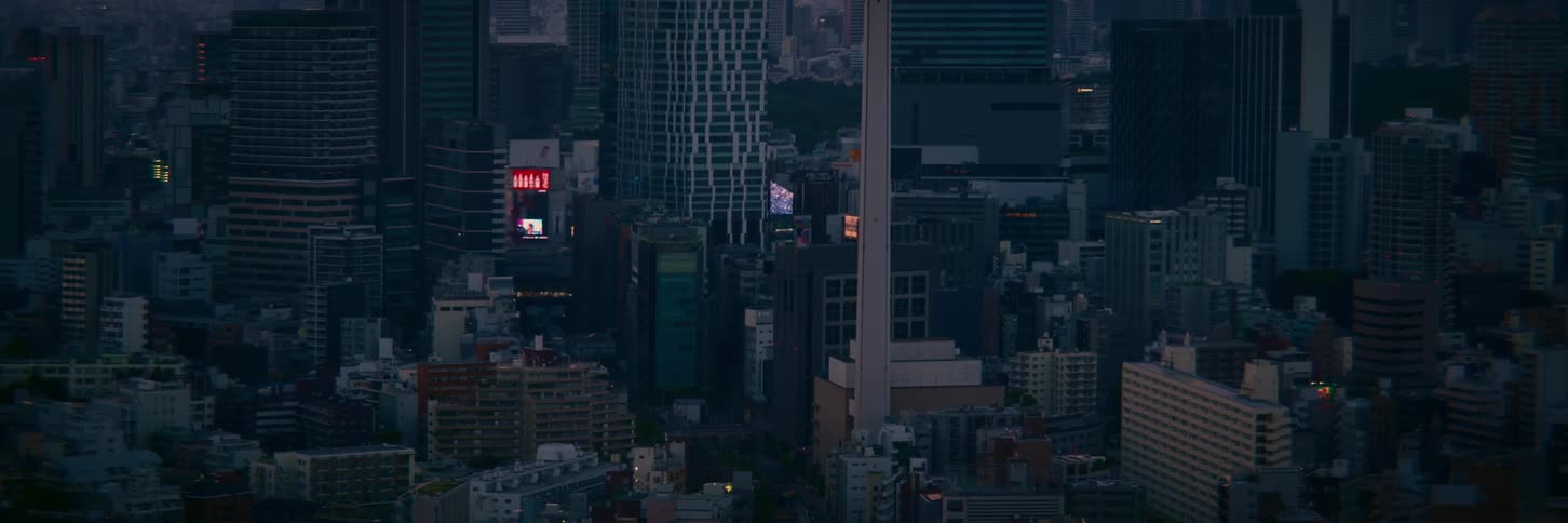A wide, static shot of a dense urban cityscape with modern architecture and illuminated billboards at dusk
