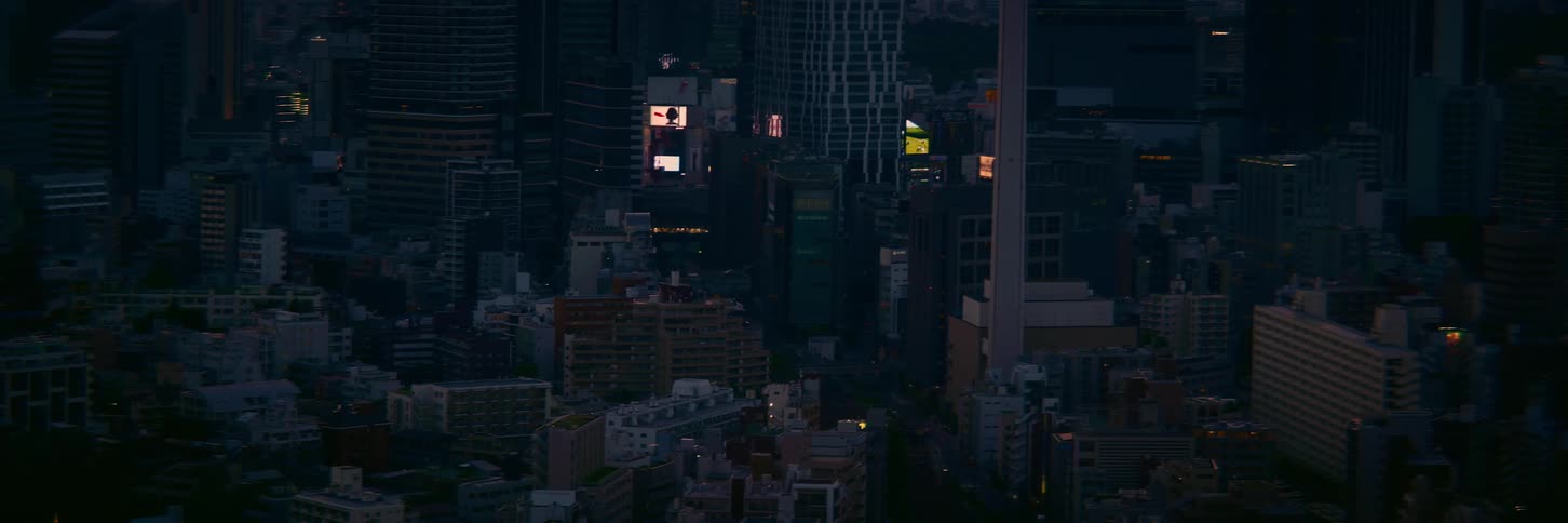 An aerial view of a dense city skyline at dusk as billboards and lights begin to illuminate the buildings