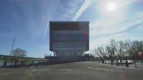 A modern, symmetrical cube-shaped building stands proudly on an empty riverside plaza on a bright and sunny day