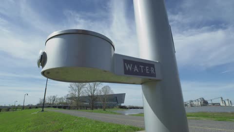 A stylish water fountain stands in a green park by the river, with an industrial backdrop under a bright blue sky