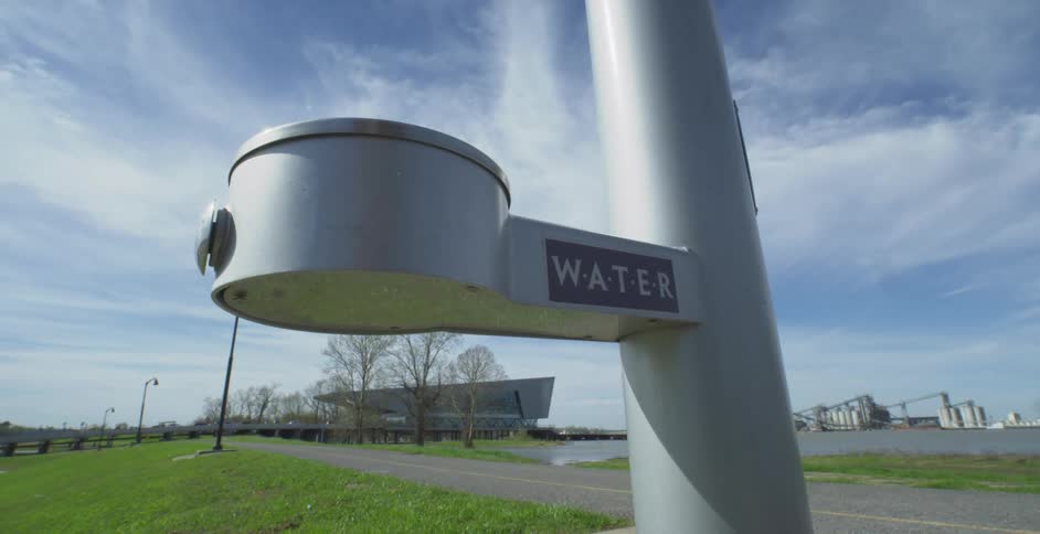 A stylish water fountain stands in a green park by the river, with an industrial backdrop under a bright blue sky