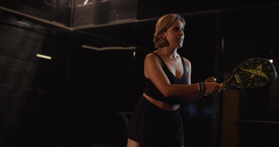A focused female paddle tennis player practices her swing in slow motion on a dark, dramatically lit indoor court