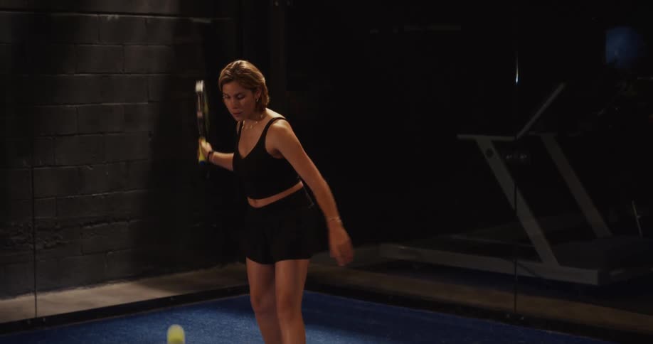A focused female athlete serves the ball in slow motion during a game of paddle tennis on a dimly lit indoor court