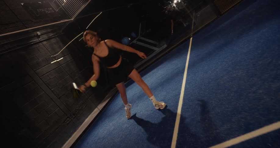 A female padel player serves the ball with a powerful swing on an indoor court, filmed in slow motion from a high Dutch angle