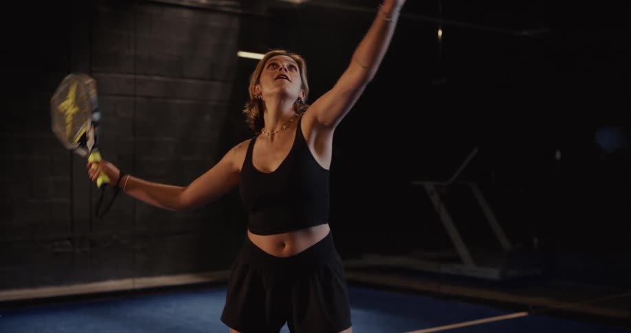A female athlete wearing black sportswear practices her serve in slow motion on a dark indoor padel court