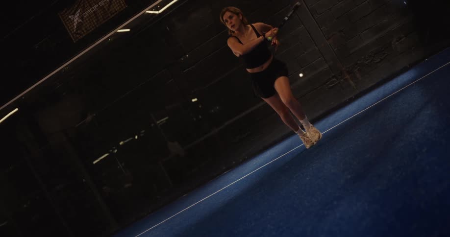 A focused female athlete plays a game of padel in dramatic slow motion on an indoor court