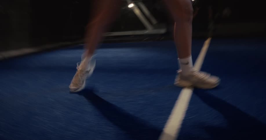 Close up low-angle view of a female athlete's fast footwork in motion blur as she plays on a blue padel court at night