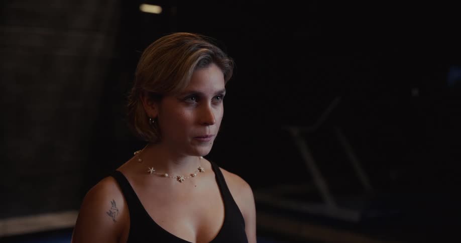 An intense female athlete catches her breath while holding a paddle ball to her head in a dark gym