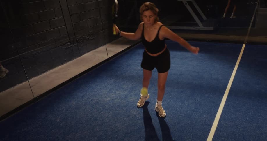 A woman serves a padel ball in slow motion on a blue indoor court
