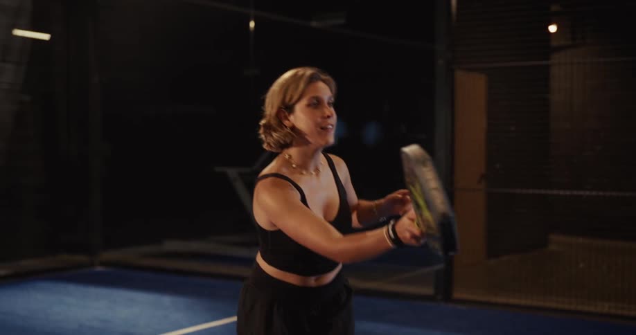 Athletic woman in black sportswear practices hitting a padel ball in slow motion on an indoor court