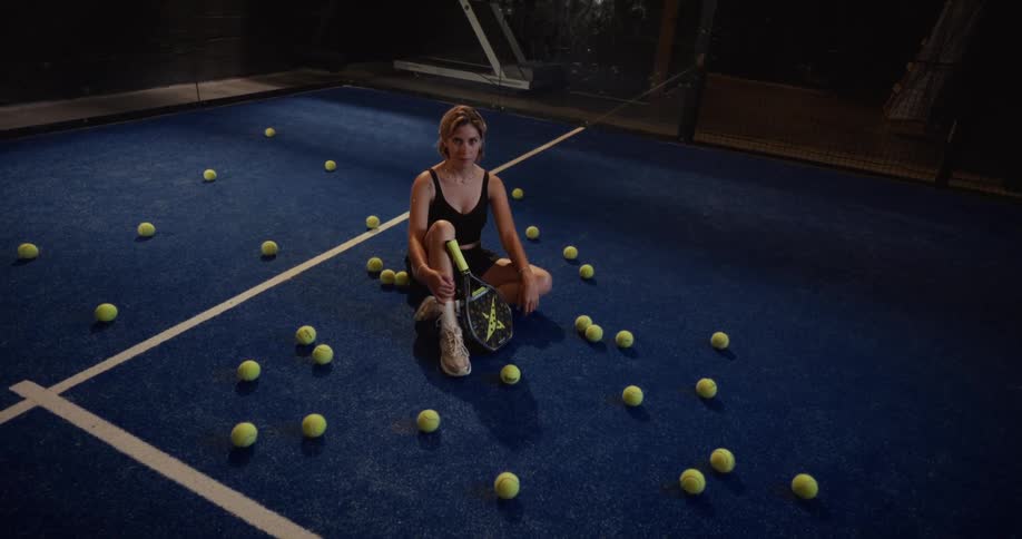 High-angle view of a fashionable woman sitting on a blue padel court at night, holding a racket and surrounded by tennis balls