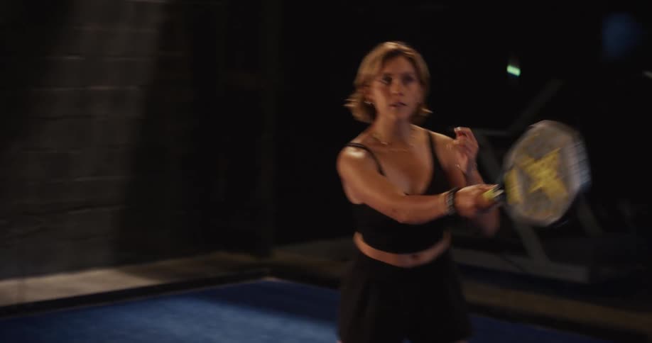 A focused young woman in athletic wear practices her padel hits in slow motion on a dark indoor court