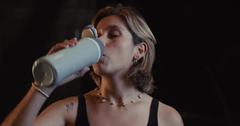 Close up of a woman taking a drink from her water bottle in slow motion in a dark gym