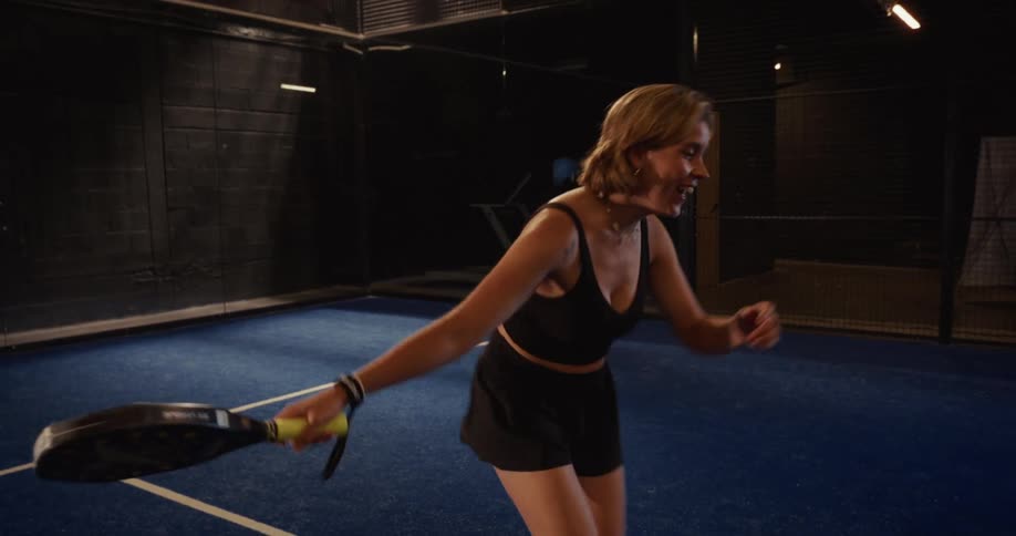 A smiling woman in black athletic wear serves a powerful overhead shot while playing padel on an indoor blue court in slow motion