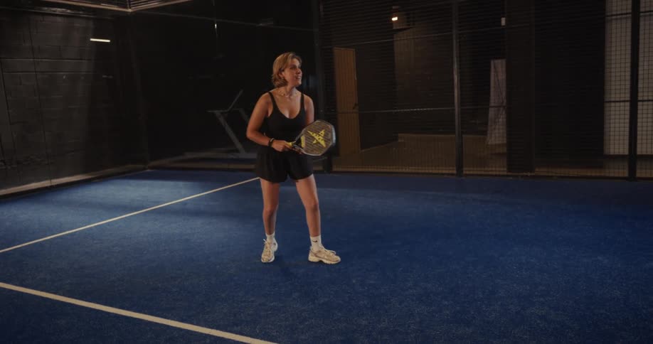 An athletic woman in black sportswear plays an energetic game of padel on a blue indoor court, captured in slow motion