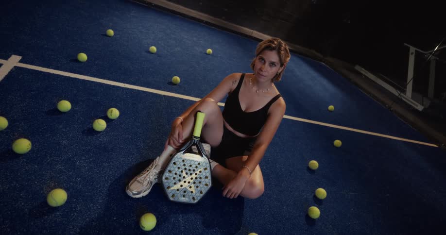 A confident female athlete sits on a blue padel court at night, holding her racket and looking directly at the camera as it slowly zooms in