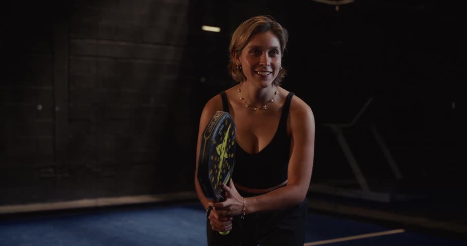 A smiling female athlete in a black sports outfit holds a racket and prepares to play paddle tennis on a dark indoor court