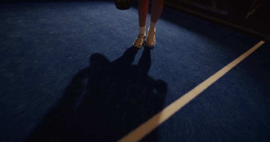 A female athlete walks backward onto a blue padel court at night, her long shadow cast by the bright overhead lights