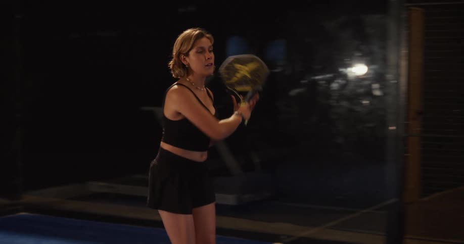 A focused woman in black athletic wear serves and plays a point during a game of padel tennis in slow motion on a dark court