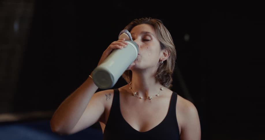 A young woman in workout clothes catches her breath and drinks water from a bottle in slow motion in a dark gym