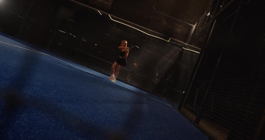 Slow motion wide shot of a woman playing padel tennis on a dark, indoor court