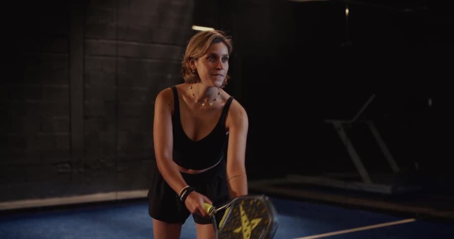 A focused female athlete serves a paddle tennis ball in slow motion on a dark indoor court