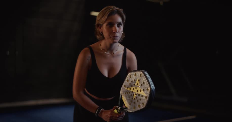 A focused female padel player stands in a ready position on a dramatically lit indoor court