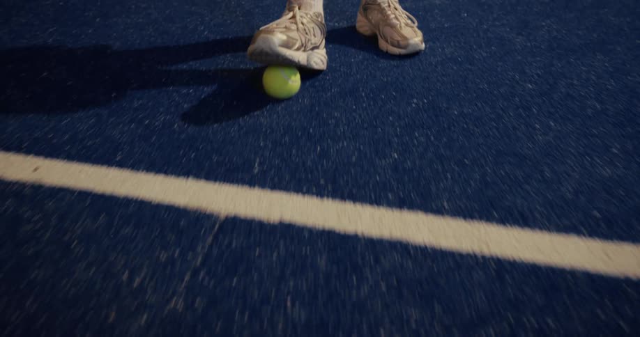 Close-up of a tennis player stopping a fast-rolling yellow tennis ball with their shoe on a blue court