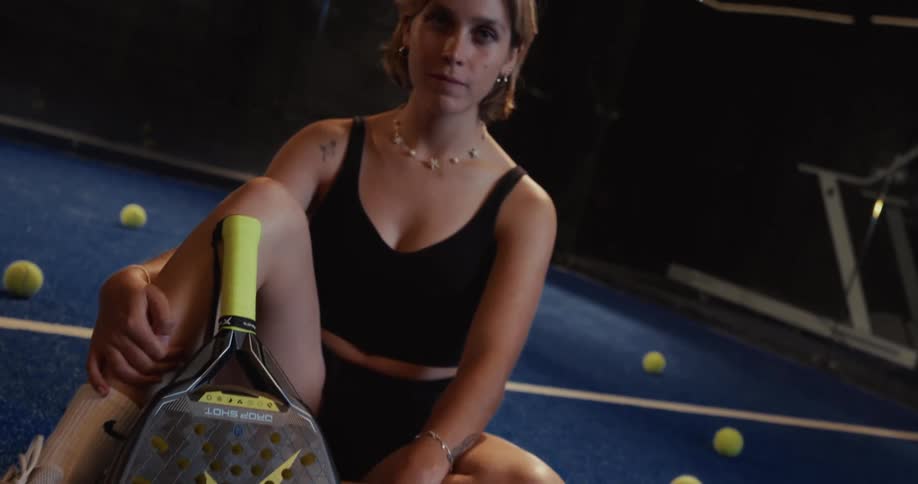 Slow motion close-up of a confident female padel player sitting on a blue court with her racket and smiling at the camera