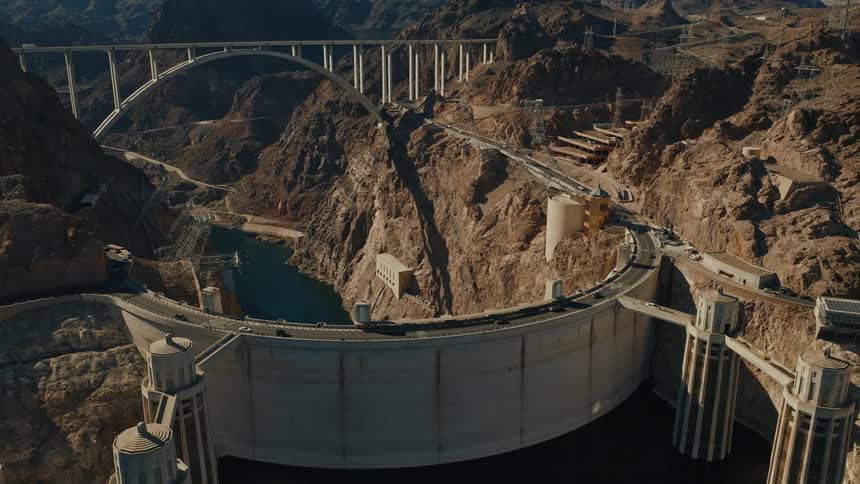 A sunny timelapse shot from a high angle shows traffic moving across the top of the iconic Hoover Dam with the Mike O'Callaghan–Pat Tillman Memorial Bridge in the background