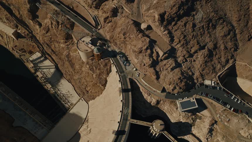 An overhead time-lapse view shows cars and people moving across the majestic Hoover Dam as shadows shift across the arid canyon