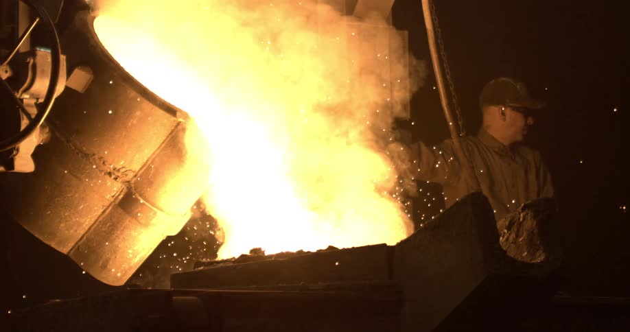 A foundry worker pours glowing molten metal into a mold in slow motion, creating a shower of sparks
