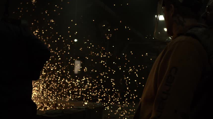 A dramatic slow-motion shot captures a brilliant shower of sparks as workers grind metal in a dark factory