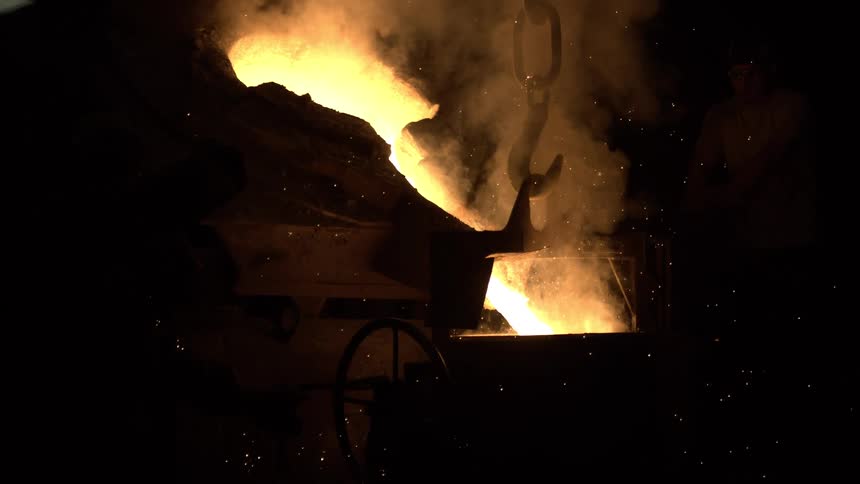 Molten metal is poured from a crucible in a dark foundry, creating smoke and sparks in slow motion