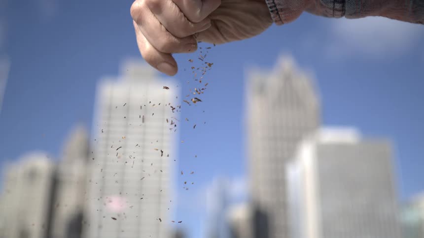 Slow motion shot of a hand sprinkling dirt with a blurred city skyline in the background
