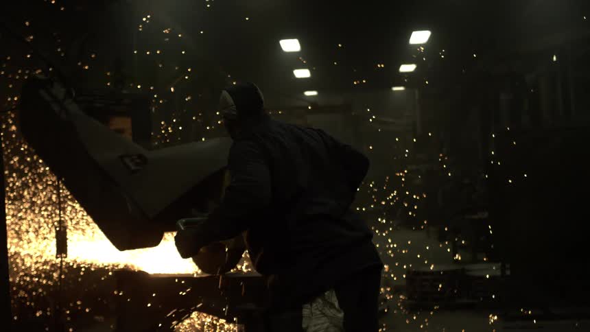 A silhouetted worker in a dark workshop uses a large industrial saw to cut metal, creating a brilliant shower of sparks in slow motion