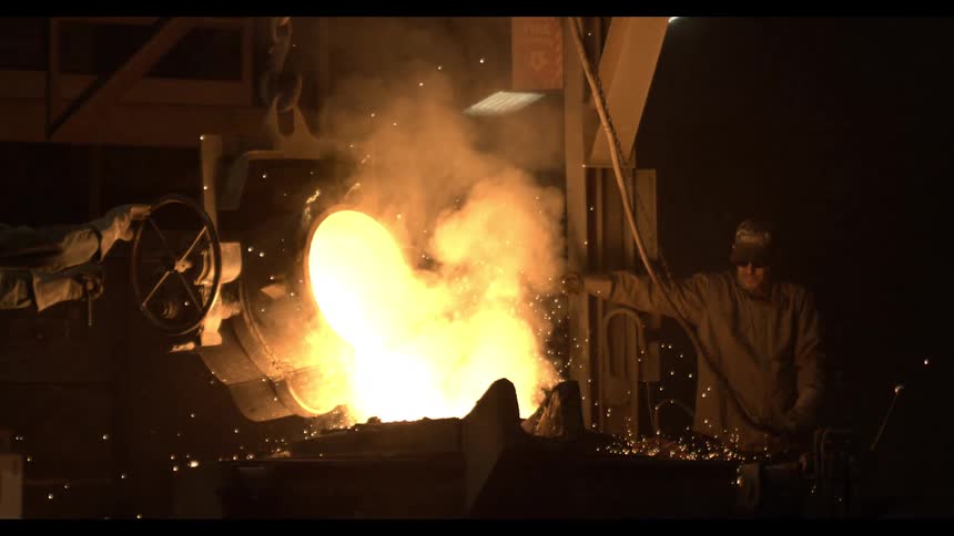 A cinematic slow-motion shot of foundry workers pouring glowing molten metal from a crucible, sending sparks flying through the dark factory