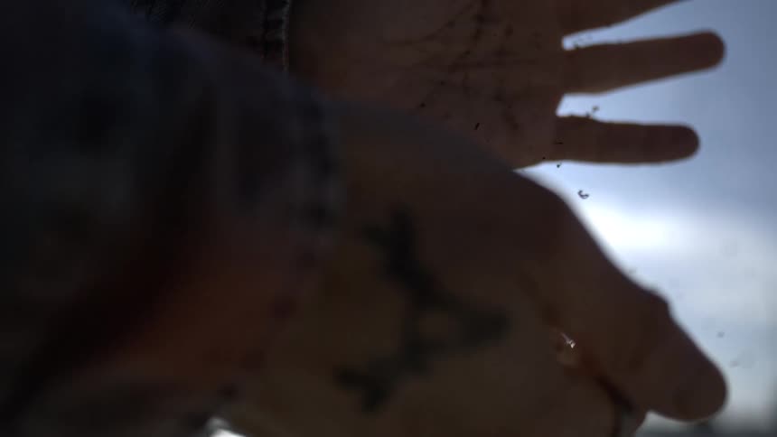 Close-up slow motion shot of loose tobacco falling through a person's tattooed hands against the bright sky