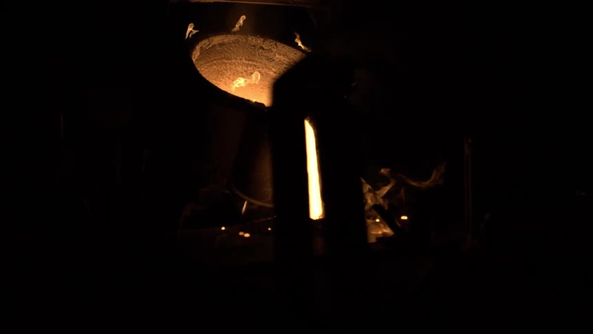 A stream of glowing hot liquid metal pours from a large crucible in a dark, smoky industrial foundry