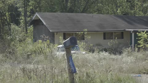 A slow zoom out of an old, abandoned rural house with an antique car parked in the overgrown front yard