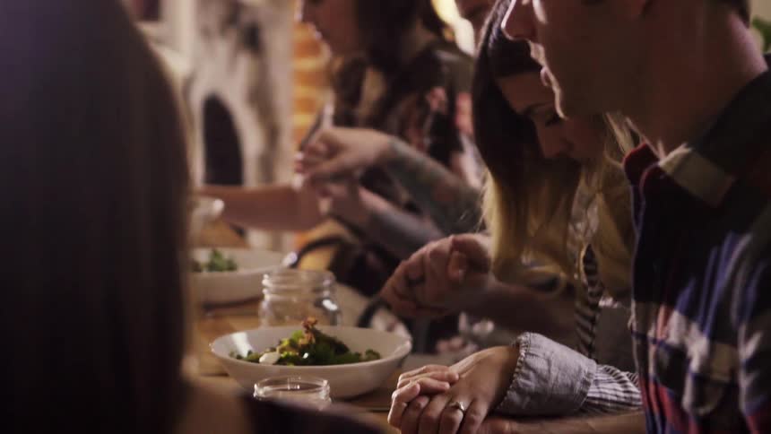 A group of dinner guests hold hands and pray before a meal