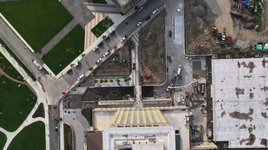 Top-down aerial view of a busy city construction site where workers pour concrete next to a finished park and active street traffic