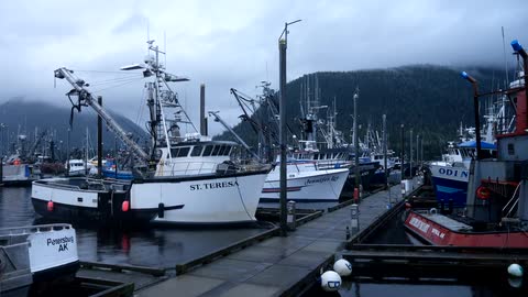 Commercial fishing boats are docked at the pier in an Alaskan harbor on a cloudy and foggy day
