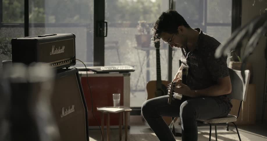 A man is practicing guitar near a large window